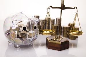 A transparent piggy bank filled with gold coins in front of stacks of gold coins, next to a gold scale of justice.